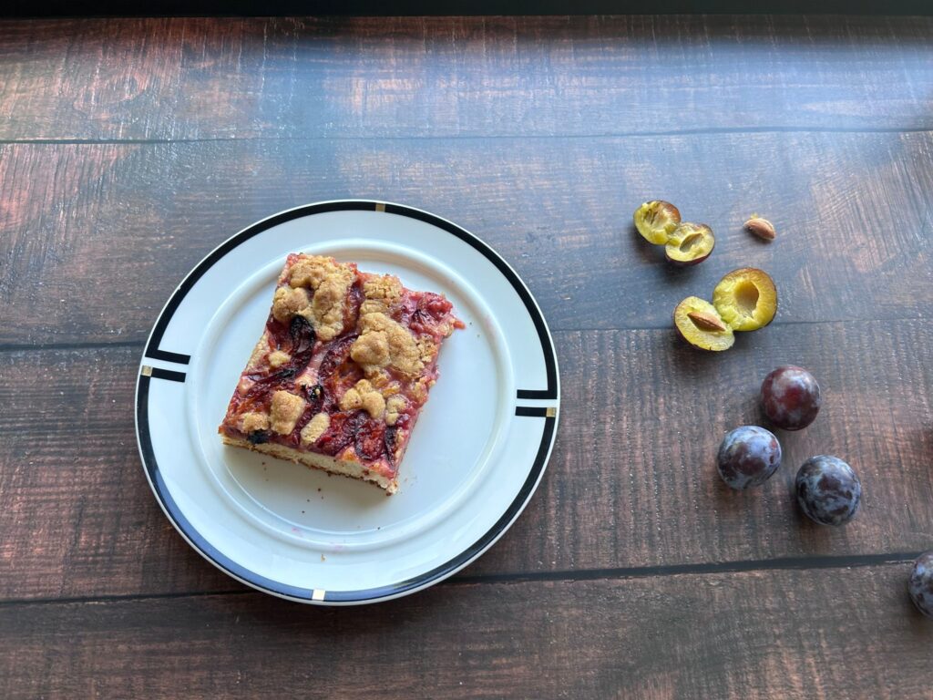 Stück Zwetschgendatschi mit Streuseln auf einem weißen Teller, daneben frische und aufgeschnittene Zwetschgen
